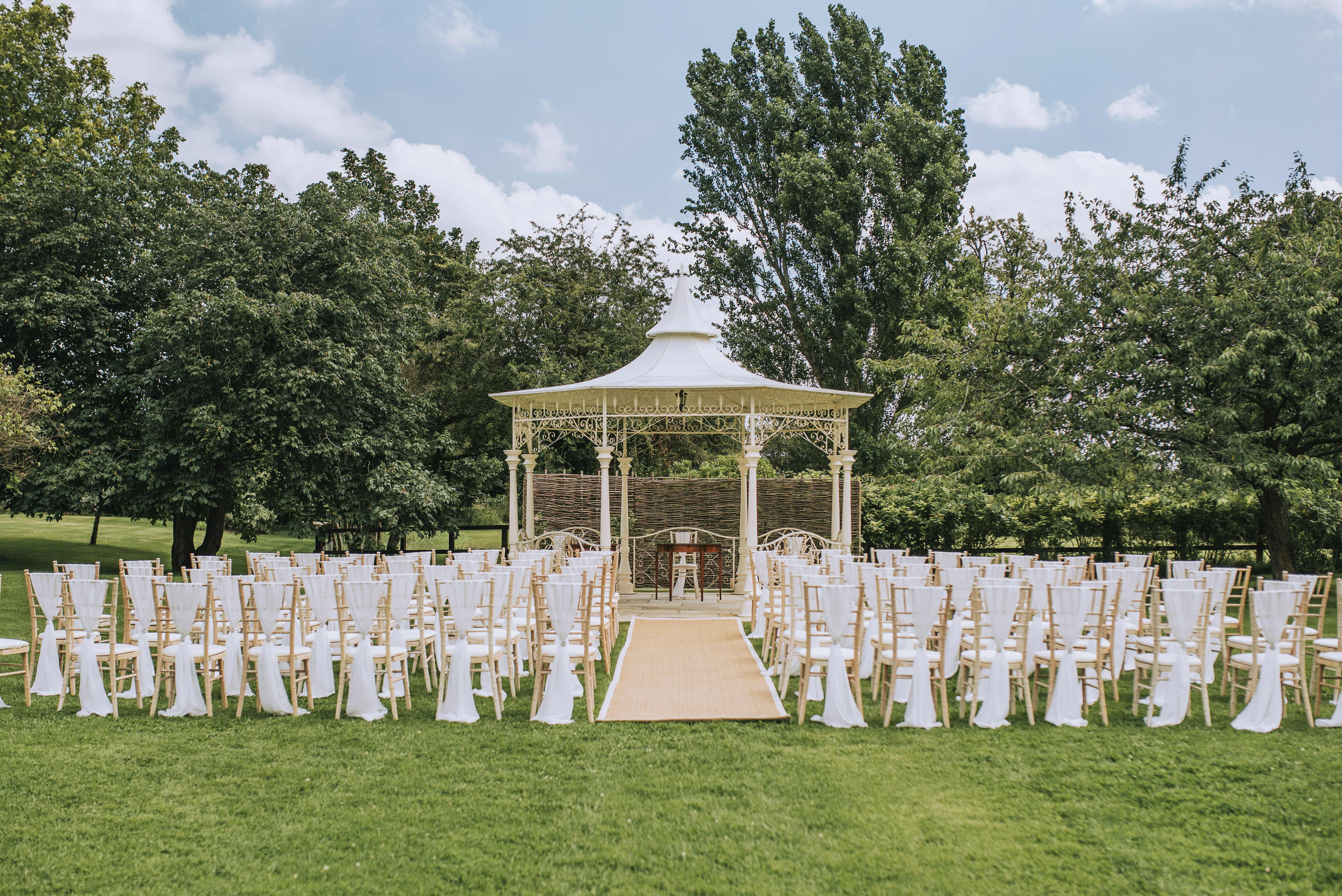 Outdoor wedding ceremony with gazebo and elegant white chairs in lush green setting.