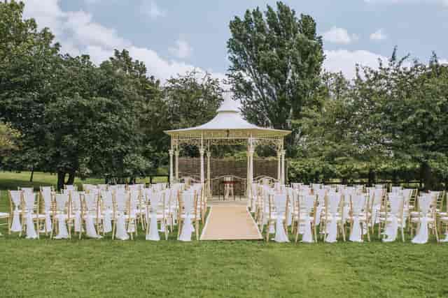 Outdoor wedding ceremony with gazebo and elegant white chairs in lush green setting.