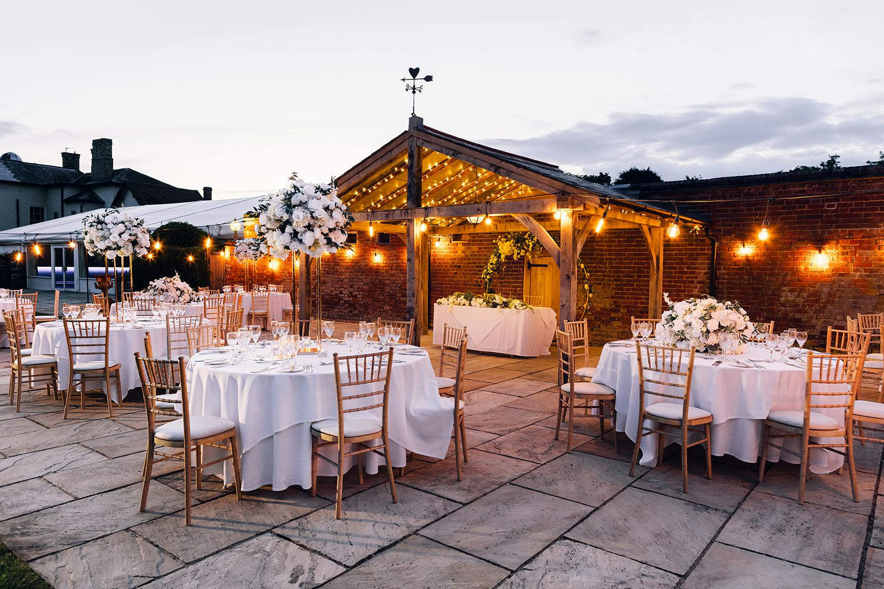 Elegant outdoor wedding venue with round tables, floral centerpieces, and string lights.