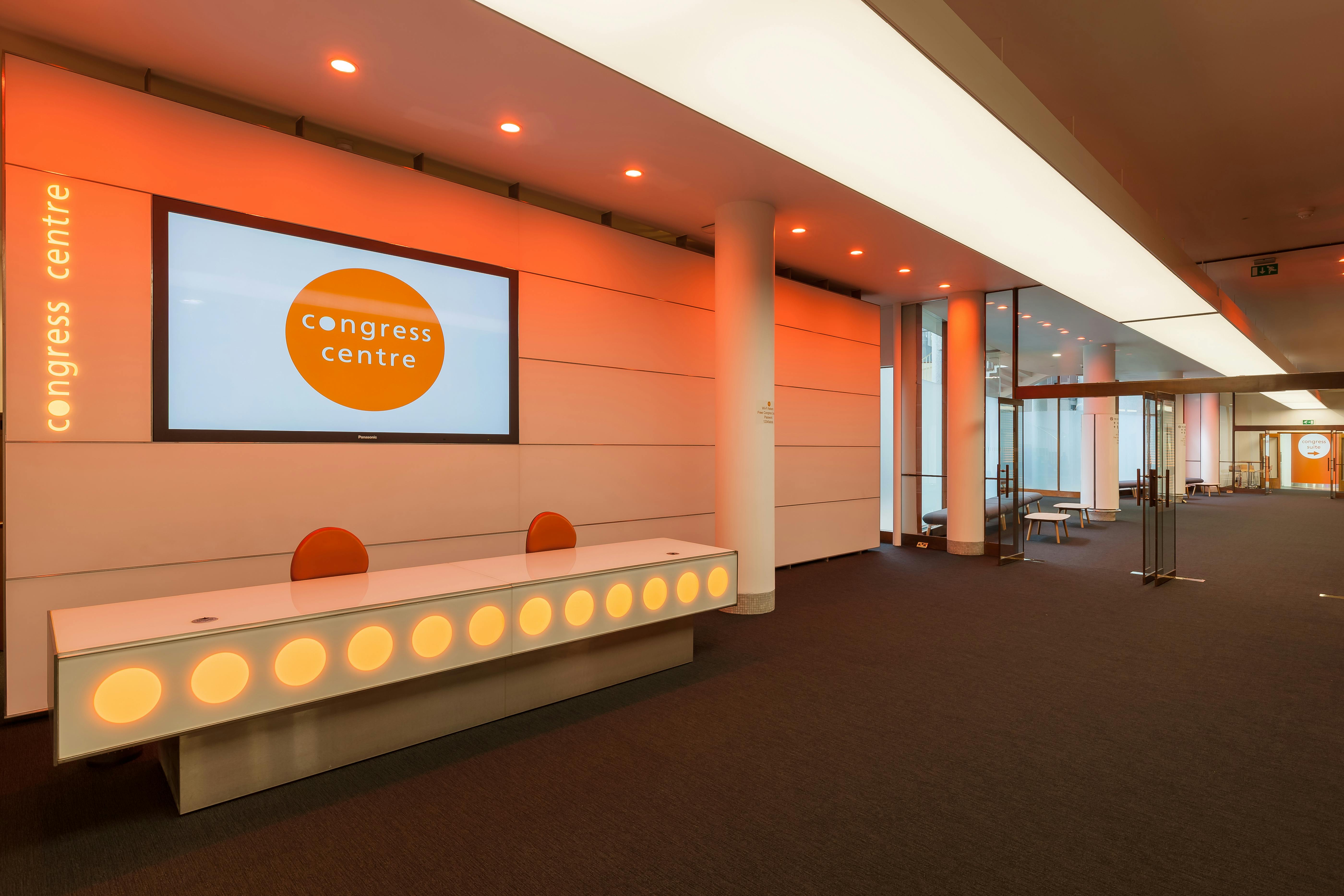 Modern congress center foyer with illuminated desk and vibrant orange accents.