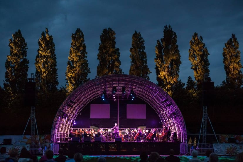 Outdoor concert at Wimbledon Park with illuminated stage and orchestra under twilight sky.