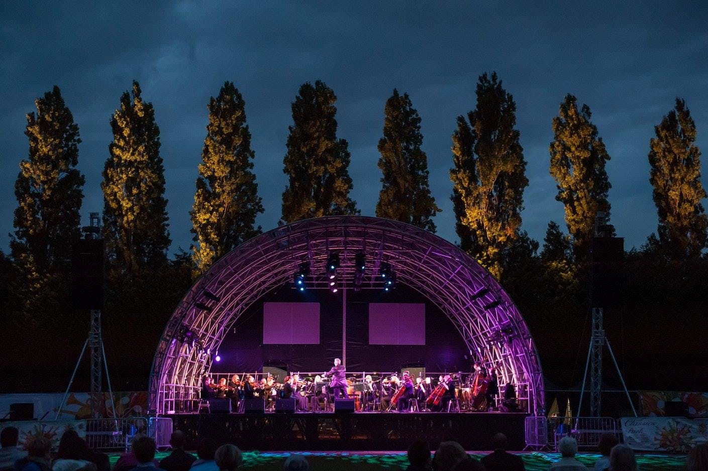 Outdoor concert at Wimbledon Park with illuminated stage and orchestra under twilight sky.