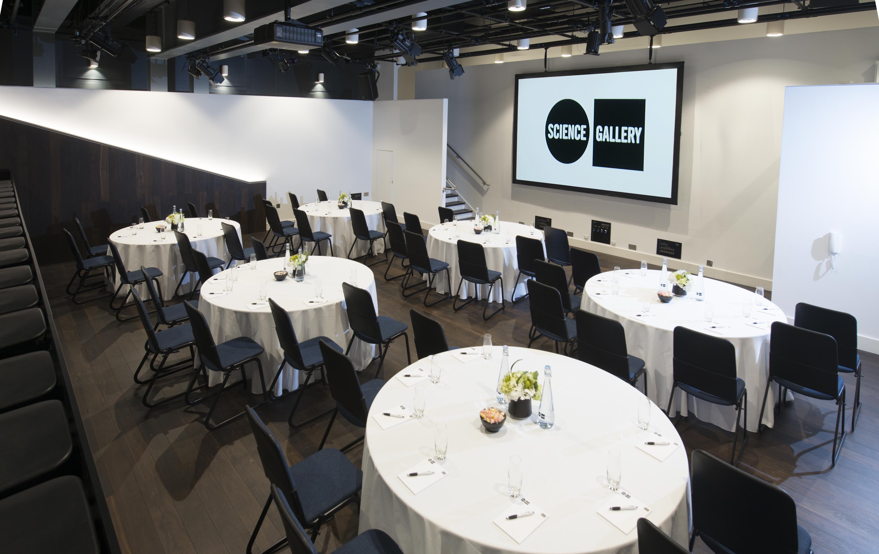 Theatre in Science Gallery London set for a modern conference with round tables and AV setup. - Banner