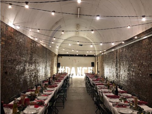 Long dining table in industrial venue for dining and dance events, illuminated by string lights. - Banner