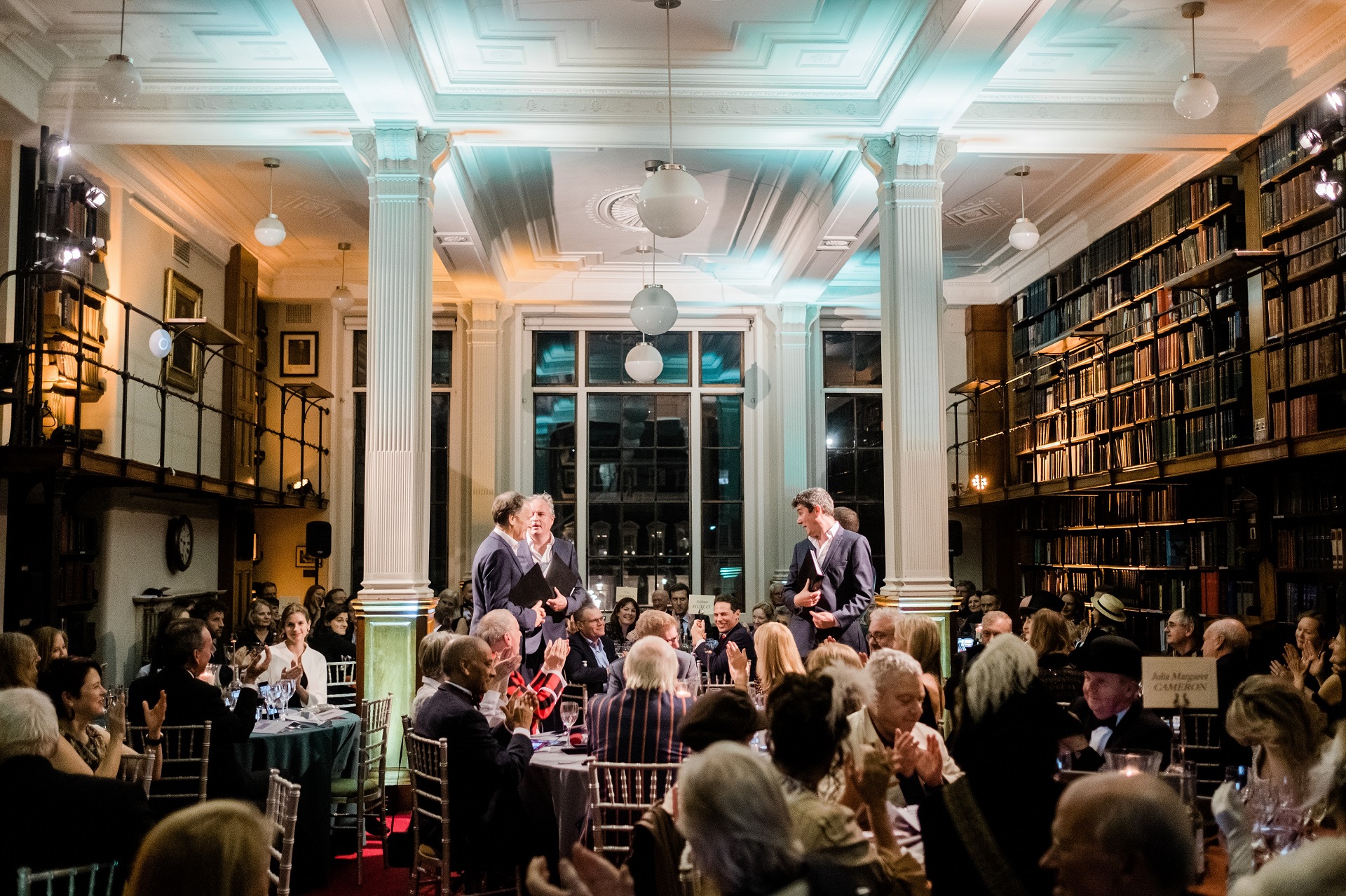 Elegant gala dinner in The London Library with guests applauding and warm lighting. - Banner