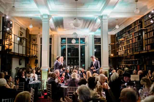 Elegant gala dinner in The London Library with guests applauding and warm lighting.