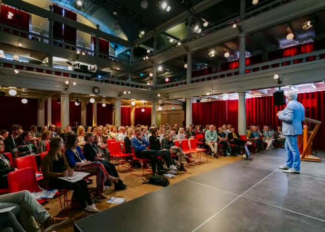 Oosterhuiszaal conference with speaker engaging audience in red chairs, ideal for networking.