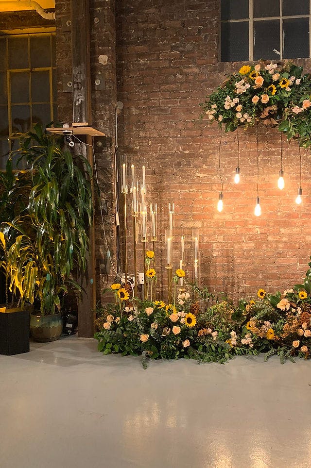 Art gallery event backdrop with rustic brick wall, floral displays, and warm lighting.