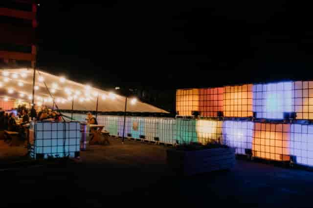 Outdoor event space with illuminated containers and string lights for gatherings.