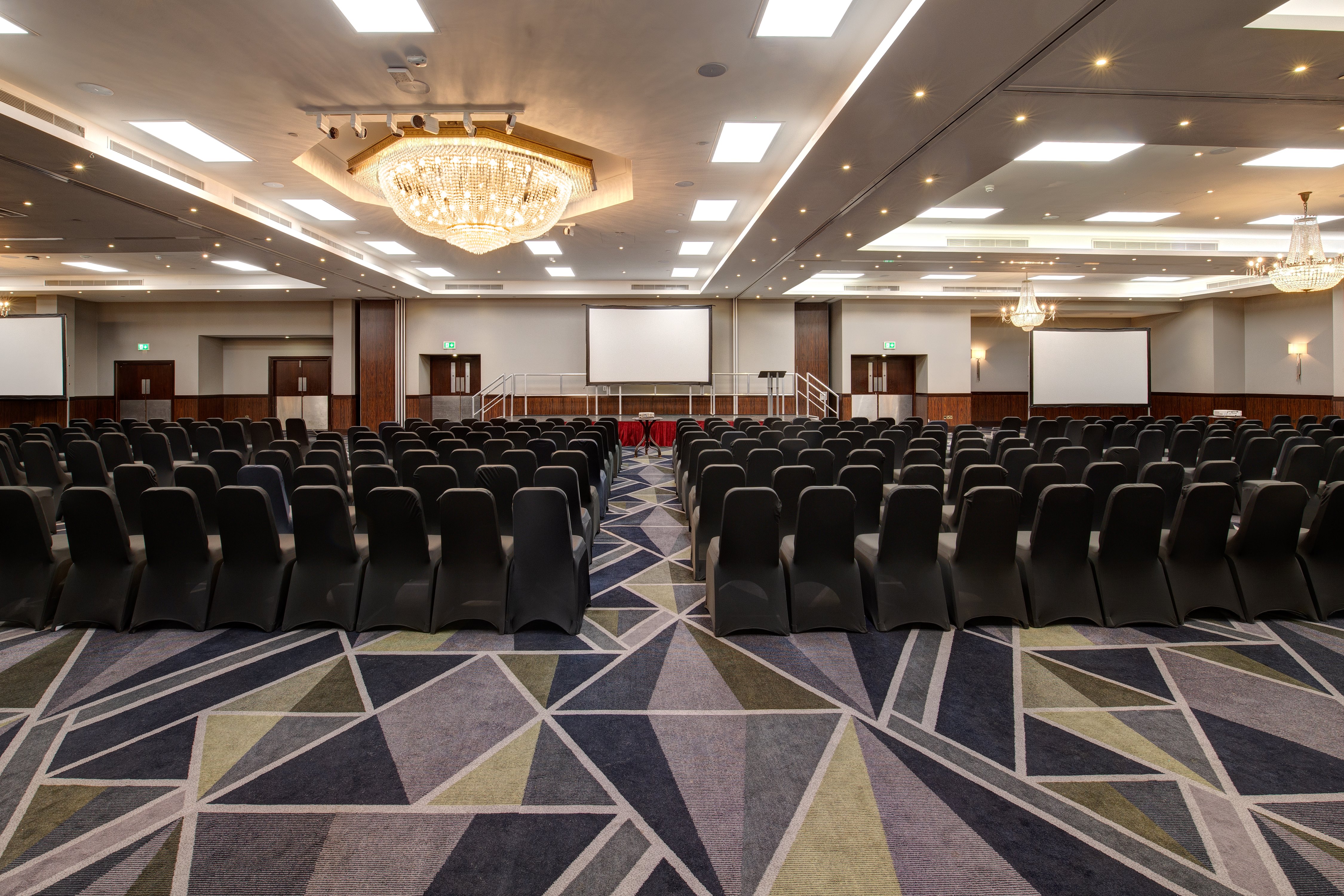 Conference room setup with black chairs and stage for presentations at Royal National Hotel. - Banner