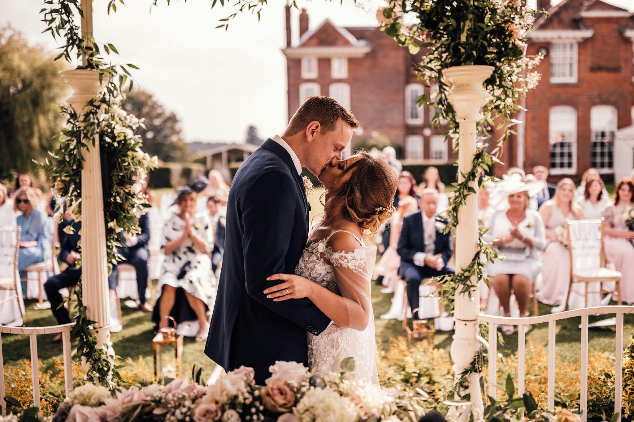 Outdoor wedding ceremony at Quendon Hall with floral arch and couple's first kiss.