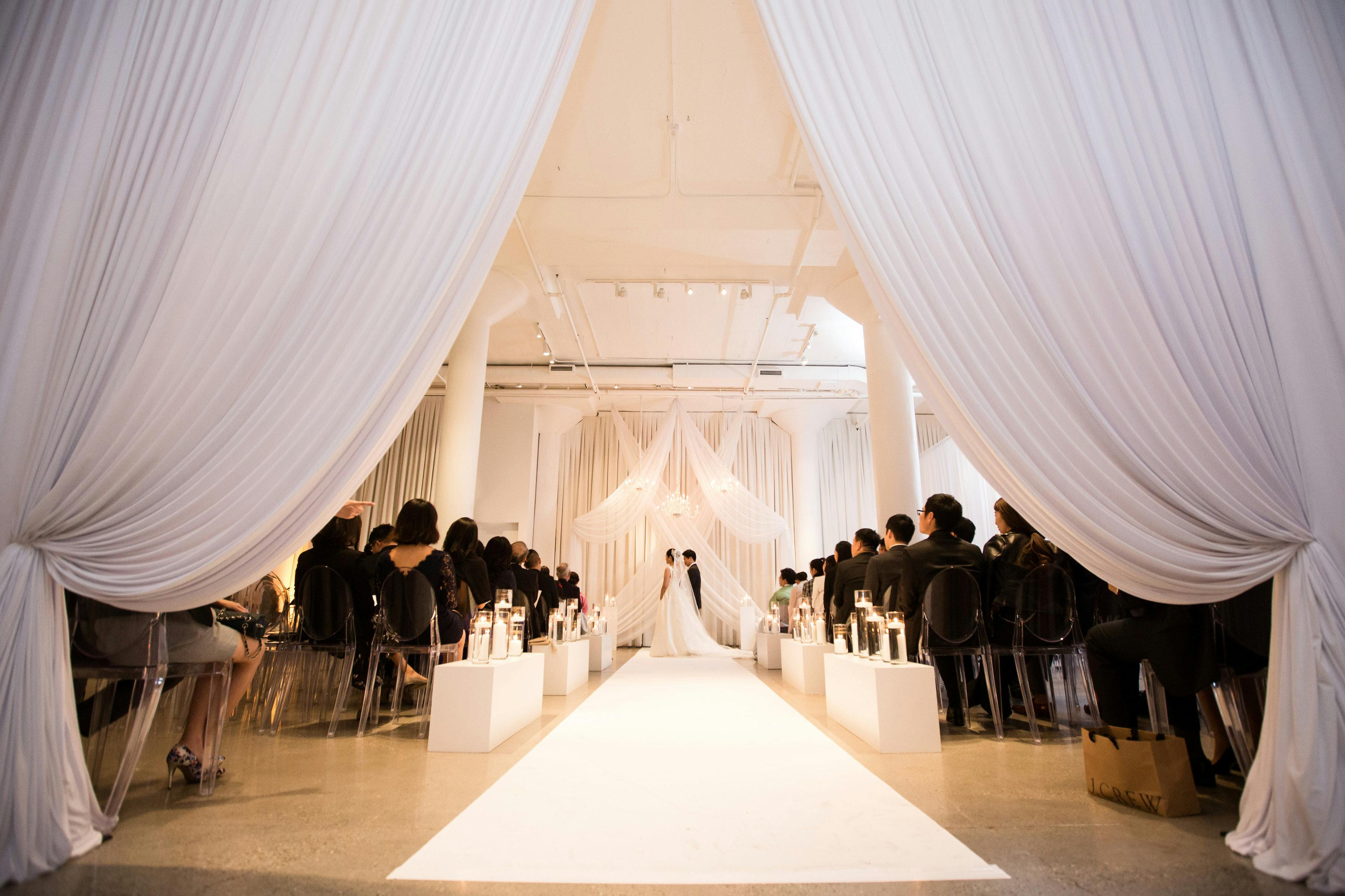 Elegant wedding ceremony in a banquet hall with white drapery and soft lighting.