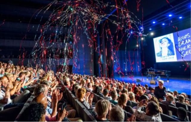 Vibrant theater event in Kunstlinie hall with enthusiastic audience and colorful streamers.