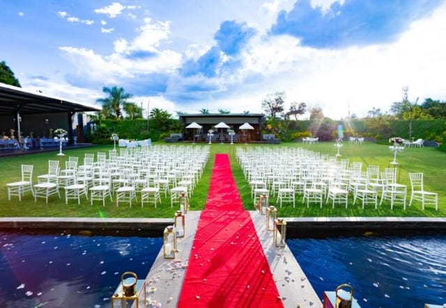Outdoor wedding venue with red carpet, elegant white chairs, and lush greenery.
