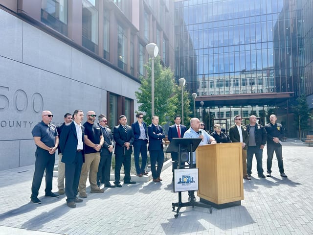"Outdoor press conference with speaker at podium, diverse audience in formal attire."