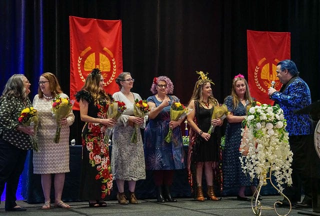 Diverse women on Flower Mound High School Theatre stage celebrate with bouquets and flags.