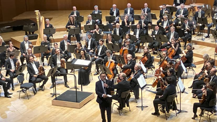 Orchestra in formal attire poised to perform at Proton de la Chapelle, Auditorium de Lyon.