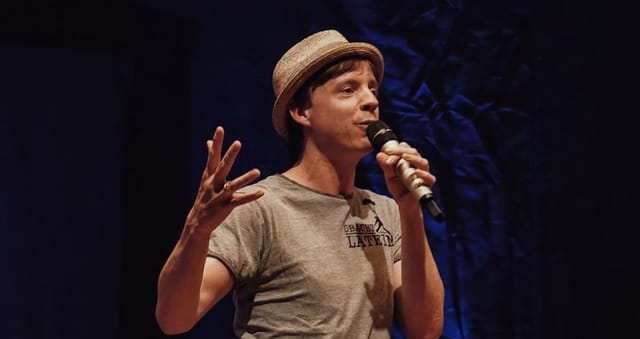 Keynote presenter at Blickpunkt Spot event with microphone against blue backdrop.