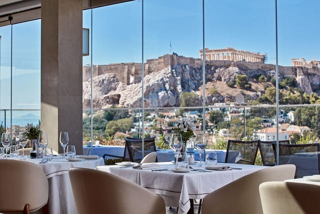 Rooftop luxury event venue with floor-to-ceiling glass and Acropolis backdrop for corporate dinners.