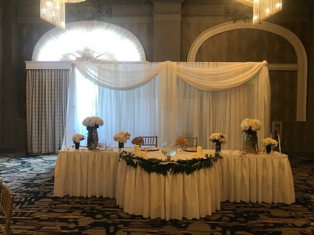 Classic wedding head table with draped backdrop and greenery for luxury banquet hall reception.