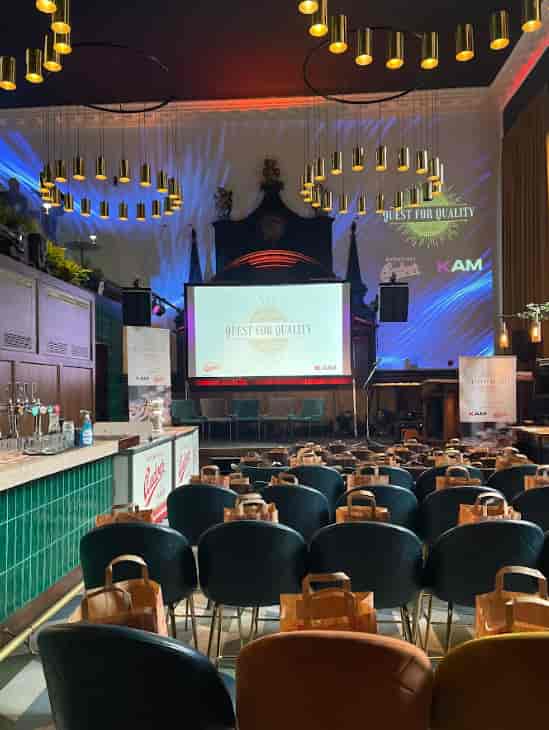 Full venue event space with green chairs and stage for "Quest for Quality" presentations.