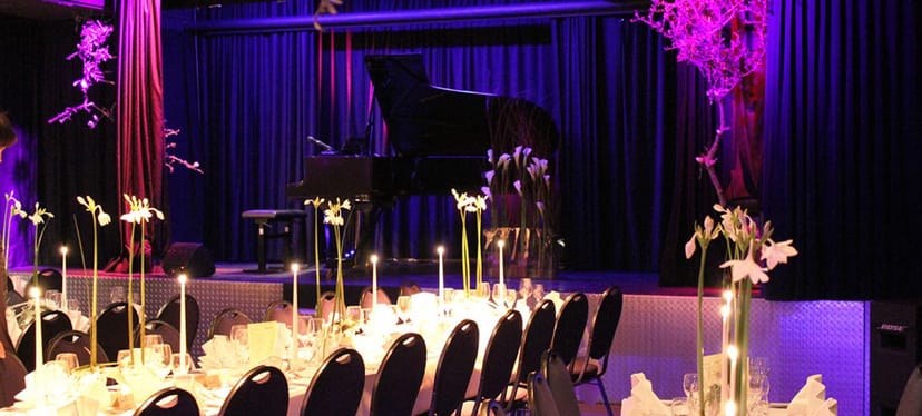Gala setup with grand piano, violet lighting, and minimalist floral decor for formal event venue.