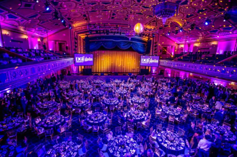 Opulent Grand Ballroom gala in Manhattan, elegantly set tables under purple lighting.