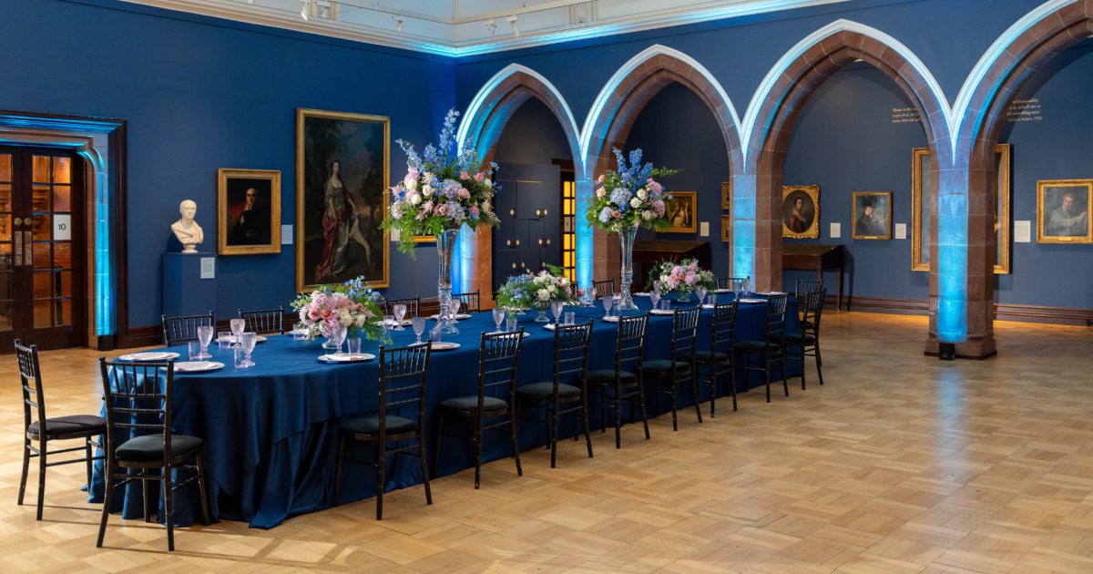 Catered gallery banquet with blue uplighting and floral centerpieces for corporate event catering. - Banner