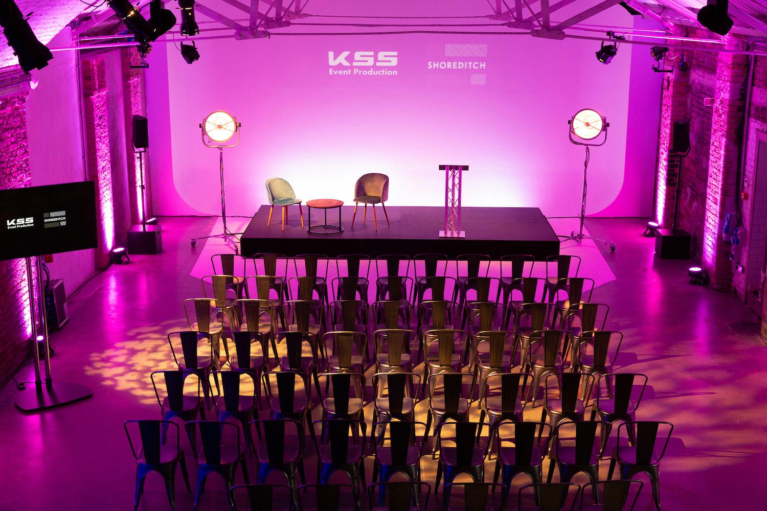 Event space in Shoreditch Studios with stage, chairs, and purple lighting for presentations.